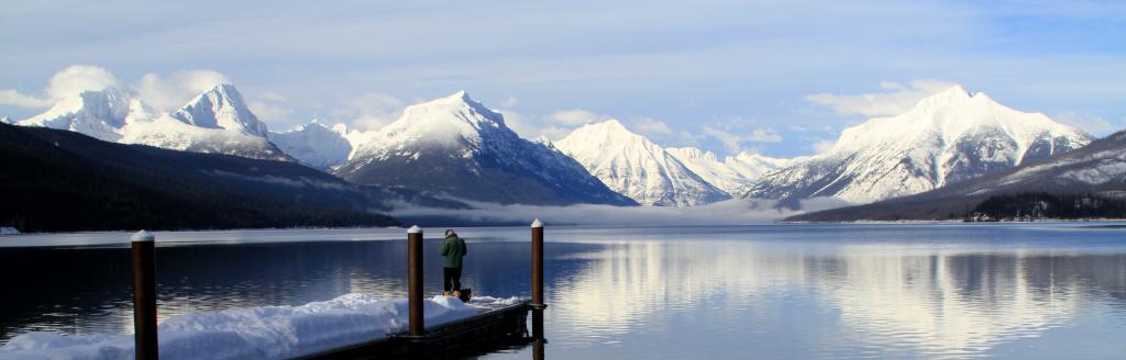 Photo. An individual standing on the end of a pier jutting into a body of water, with snowcapped mountains in the distance.